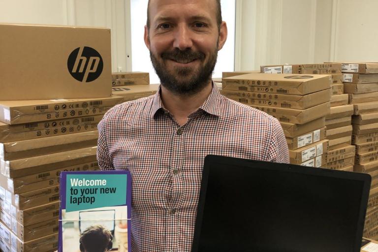 A lambeth councillor holding a laptop and a 'welcome to your new laptop' leaflet with lots of laptops in boxes behind them