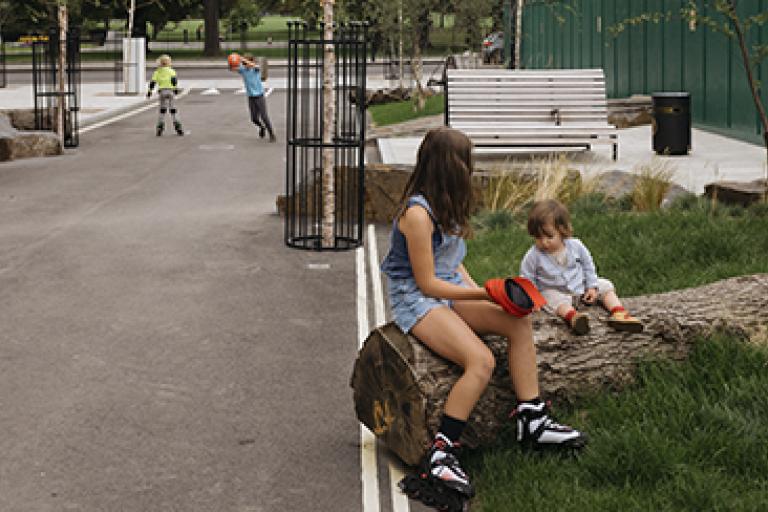 a child and a toddler sat on a log which is on a street that has been designed to encourage walking and playing