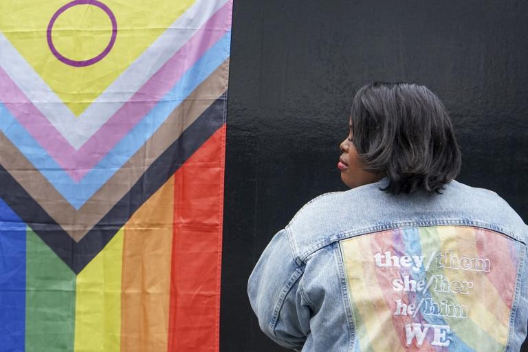 Lady in jean jacket faces backwards standing next to pride flag