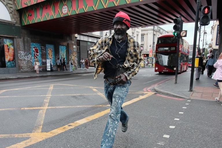Photo of man in red wool hat and camouflage jacket running on pavement with Brixton railway bridge behind him
