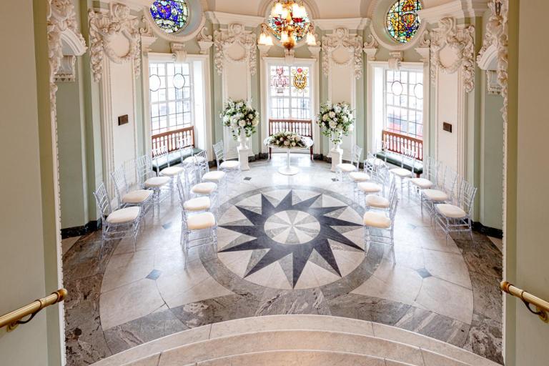 Wedding ceremony room at Lambeth Town Hall with stained glass windows, floral displays and chairs arranged around a patterned marble floor