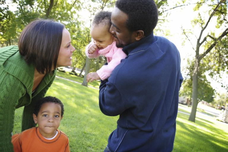 Two parents, one parent holding a baby, with the other parent looking at the baby. There is another child stood with the parent.