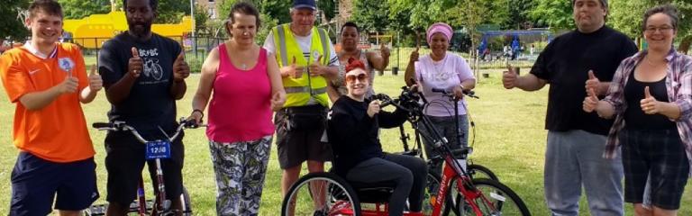 A range of different people posing for a photo in a park holding their thumbs up