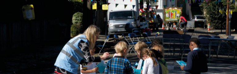 A woman bending down to talk to some children who are playing on a 'play street' a road closed to traffic to allow kids to play.