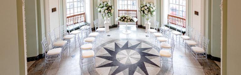 Wedding ceremony room at Lambeth Town Hall with stained glass windows, floral displays and chairs arranged around a patterned marble floor