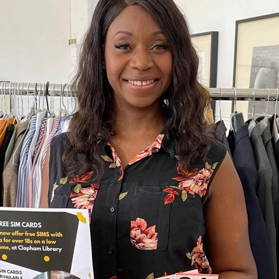 Cllr Manley-Browne holds SIM cards and poster with details of the free SIM card scheme from Lambeth libraries. She is standing in front of a rail of donated smart & designer clothes that residents can borrow from the library for job interviews.