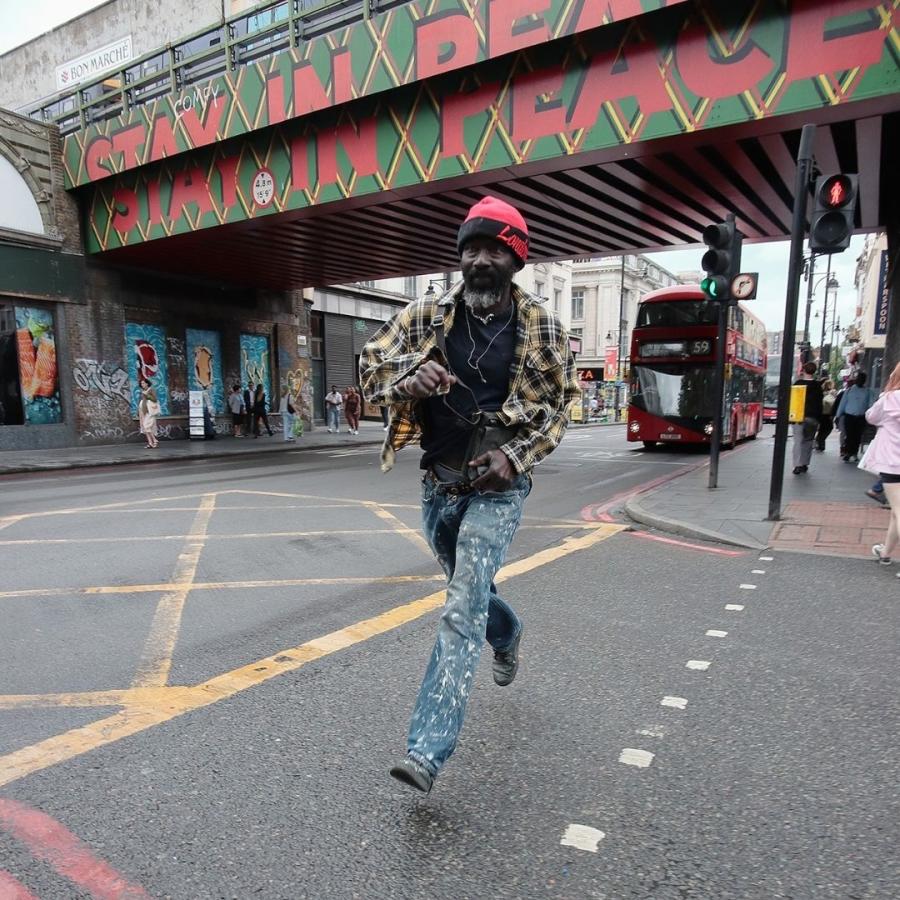 Photo of man in red wool hat and camouflage jacket running on pavement with Brixton railway bridge behind him