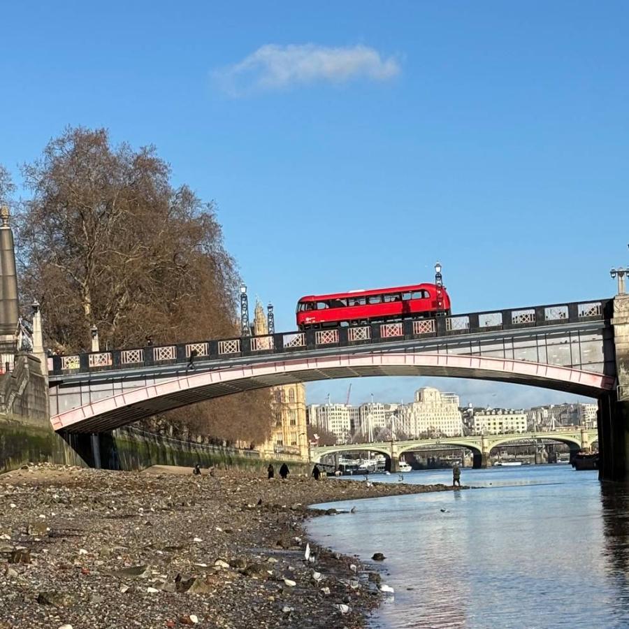 Red bus crosses bridge above Thames beneath bright blue London sky.