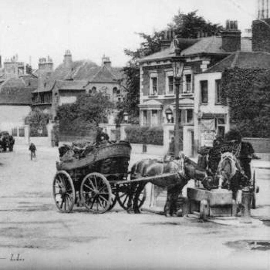 Historic Clapham street with horse-drawn carts, cyclists, pedestrians beside buildings.