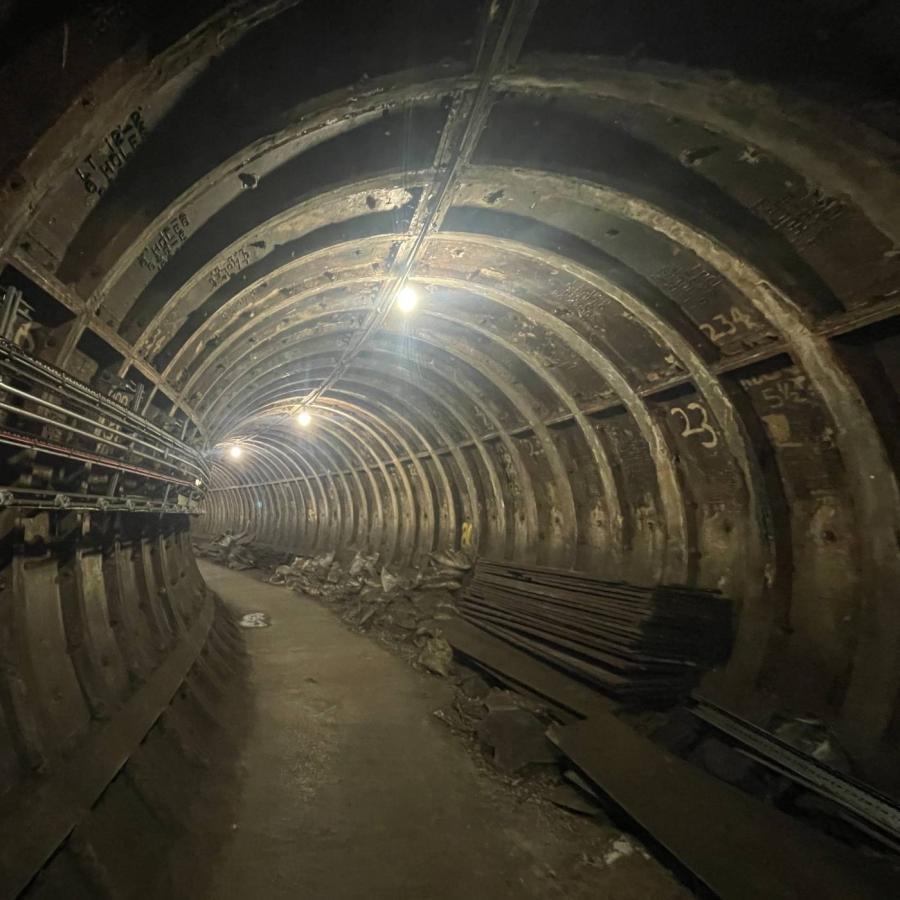A dimly lit industrial tunnel curves into darkness, lined with metal ribs, exposed cables, scattered debris, stacked materials, and overhead lights.