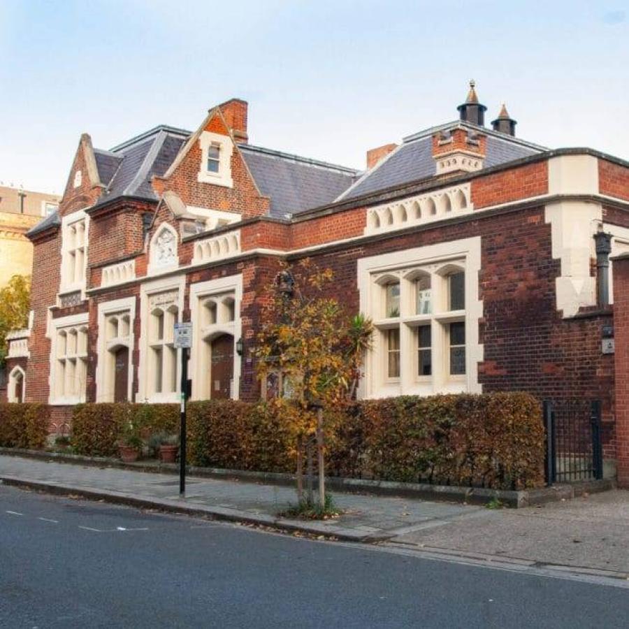 A stately red-brick heritage building with ornate stone detailing, steep slate roofs, and large mullioned windows, fronted by neatly trimmed hedges along a quiet street.