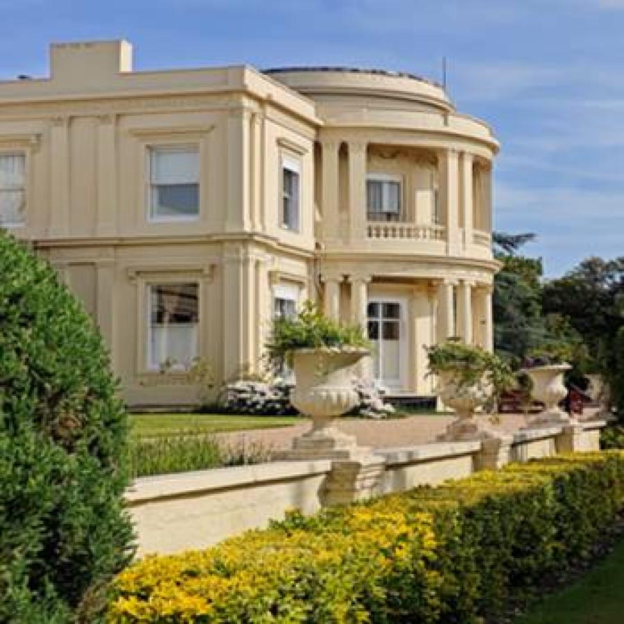 Elegant cream-colored mansion surrounded by manicured gardens, hedges, decorative planters, under blue sky.