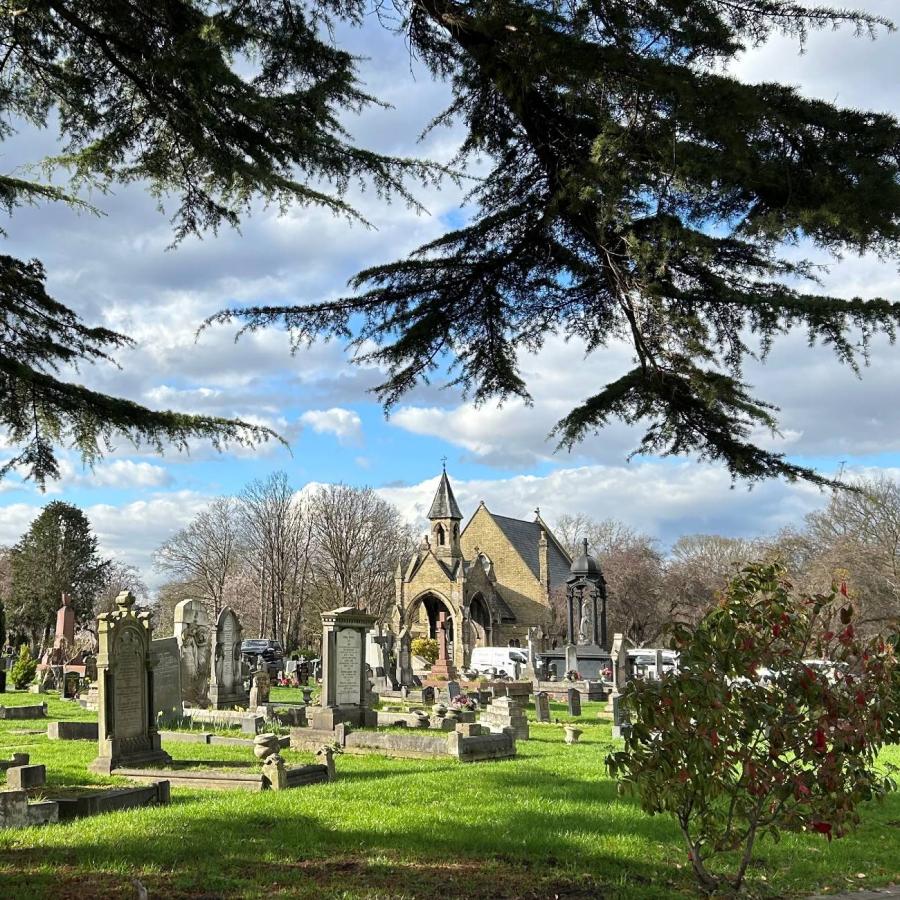 Peaceful cemetery with historic chapel, gravestones, towering trees, and cloudy blue sky