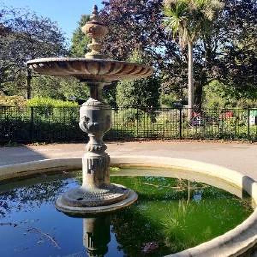 Stone fountain reflecting greenery beside pathways in a sunny park.