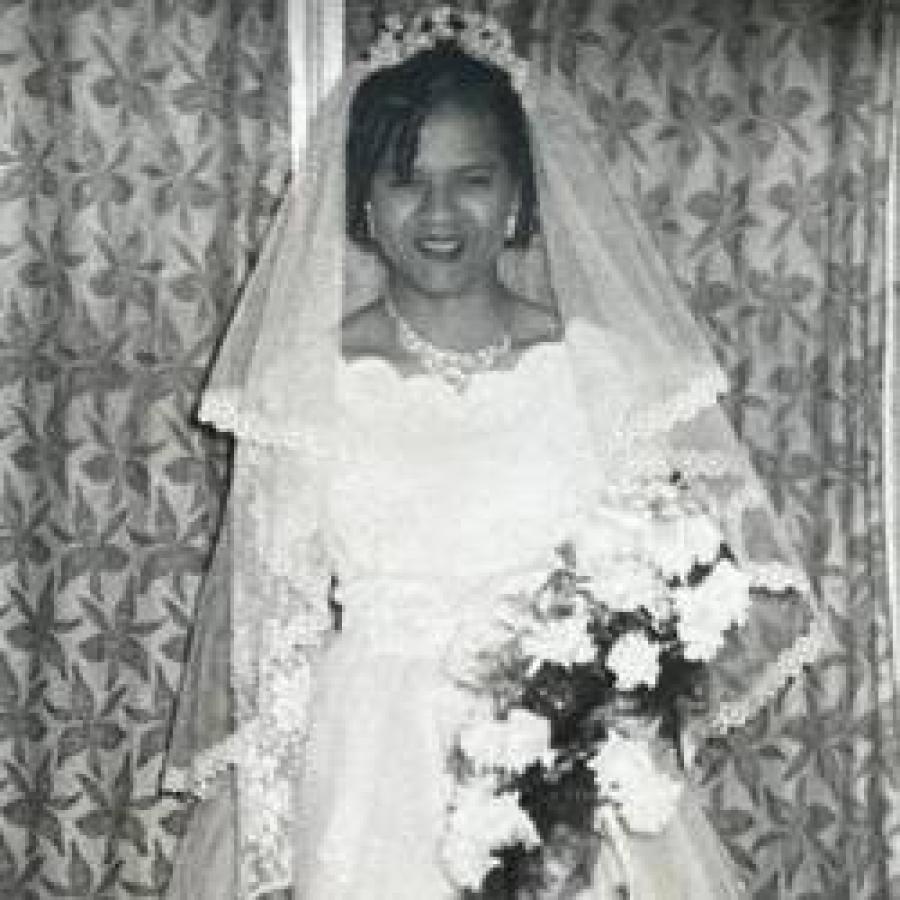 Bride in vintage wedding dress holding bouquet, smiling beside patterned curtains.